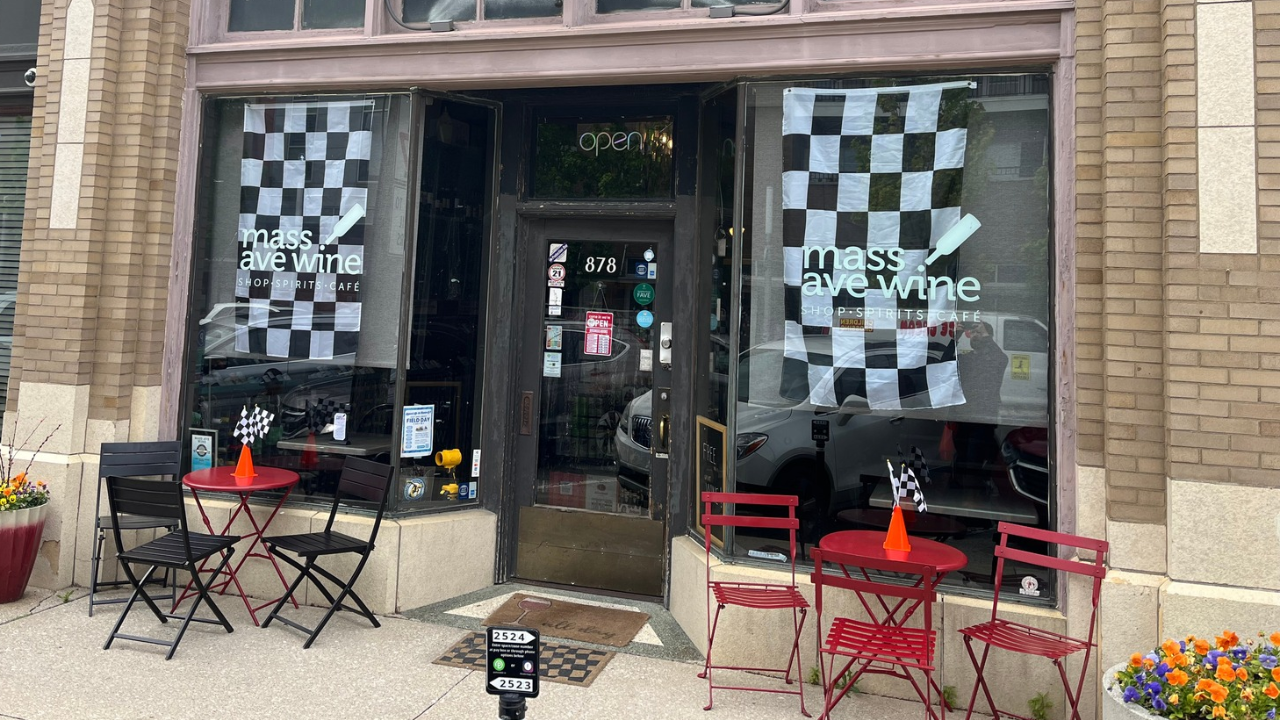 Mass Ave Wine shop's exterior with checkered flags on bistro tables inside a small yellow traffic cone. Two large flags appear hanging in the front windows.